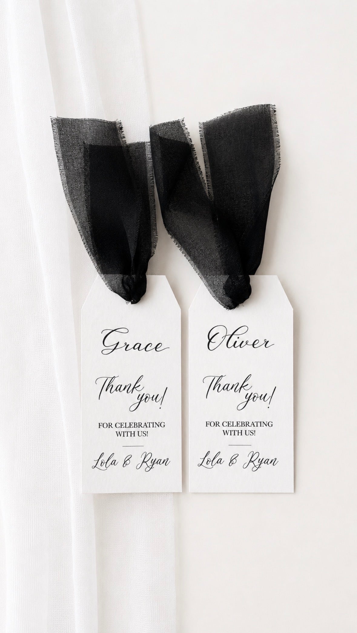 Two white personalised party favor tags with black ribbon bows, handwritten with names and thank you messages, hanging against a white curtain background.