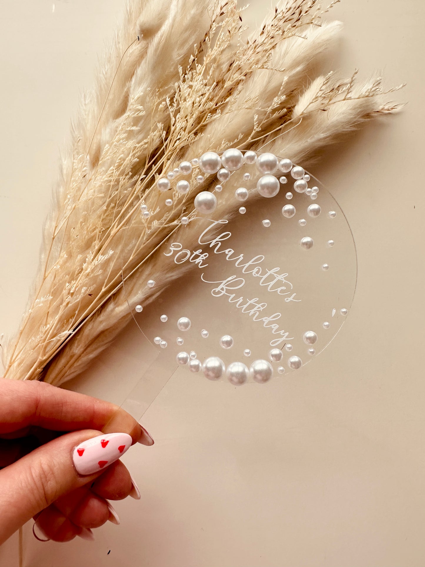 A clear acrylic cake topper with white pearl detailing and elegant calligraphy text, displayed on a backdrop of soft, neutral-toned pampas grass. The topper is personalised with a custom name and adds a luxurious, modern touch to any wedding or celebration cake.