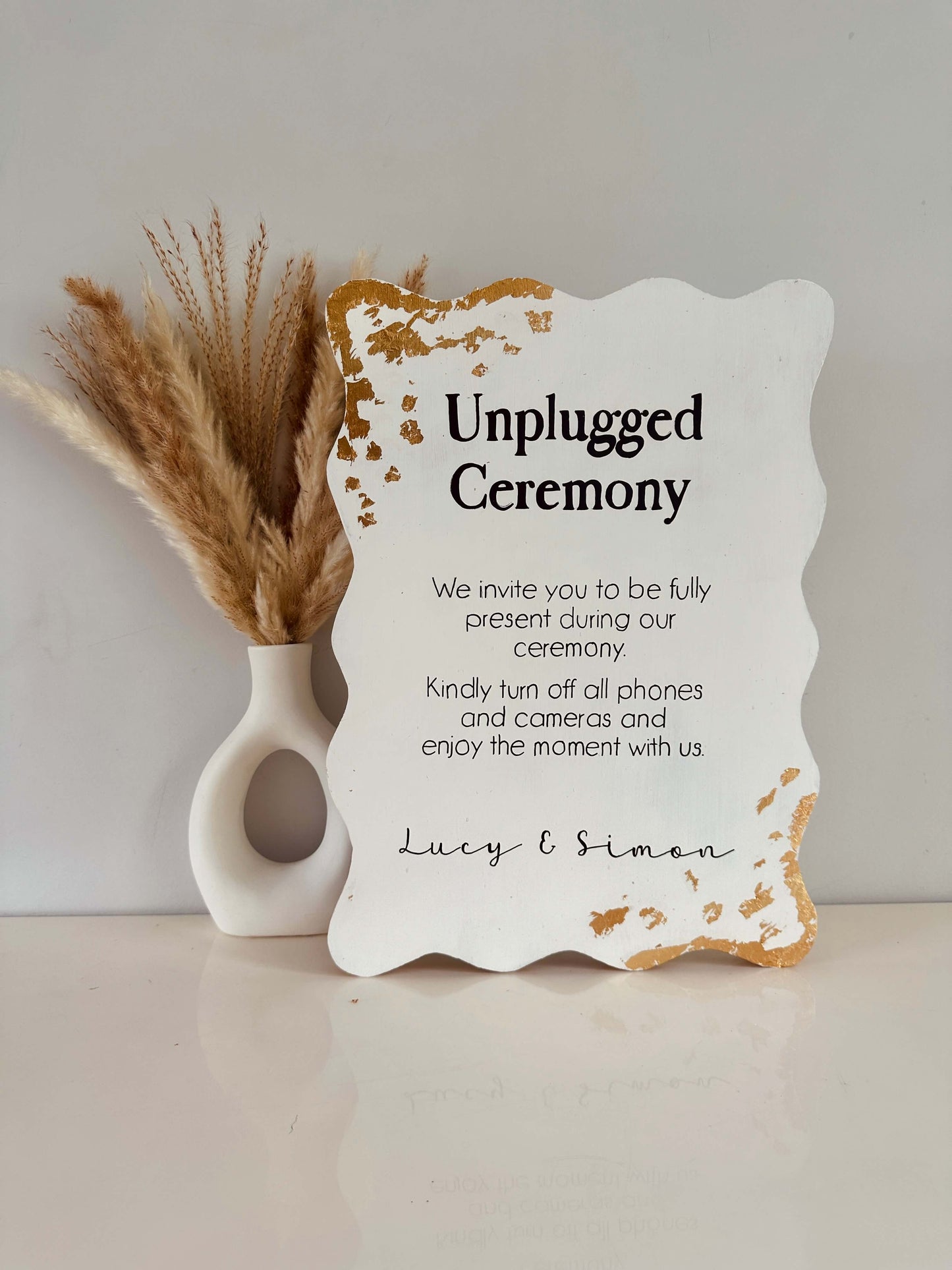 Unplugged ceremony sign with decorative vase on a neutral background. This freestanding 18mm thick wooden sign is finished with black writing reading unplugged ceremony' with gold flakes in the corners for extra sparkle