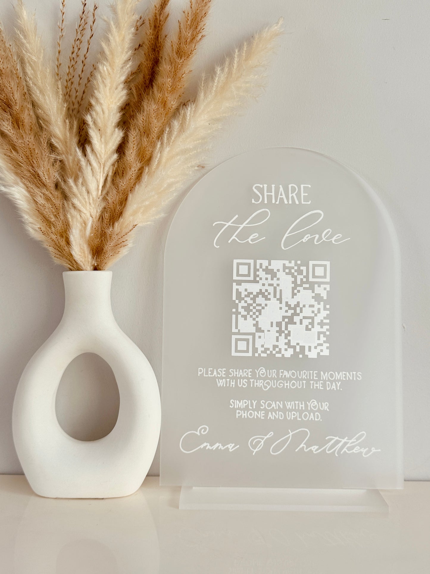 White vase with dried pampas grass and a clear acrylic arch with QR code on with white writing saying 'share the love, please share your favourite moments with us throughout the day. Simply scan with your phone and upload' finished with couples names at the bottom.