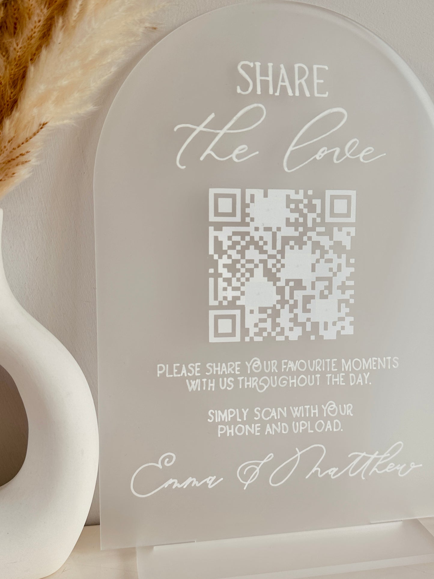 White vase with dried pampas grass and a clear acrylic arch with QR code on with white writing saying 'share the love, please share your favourite moments with us throughout the day. Simply scan with your phone and upload' finished with couples names at the bottom.