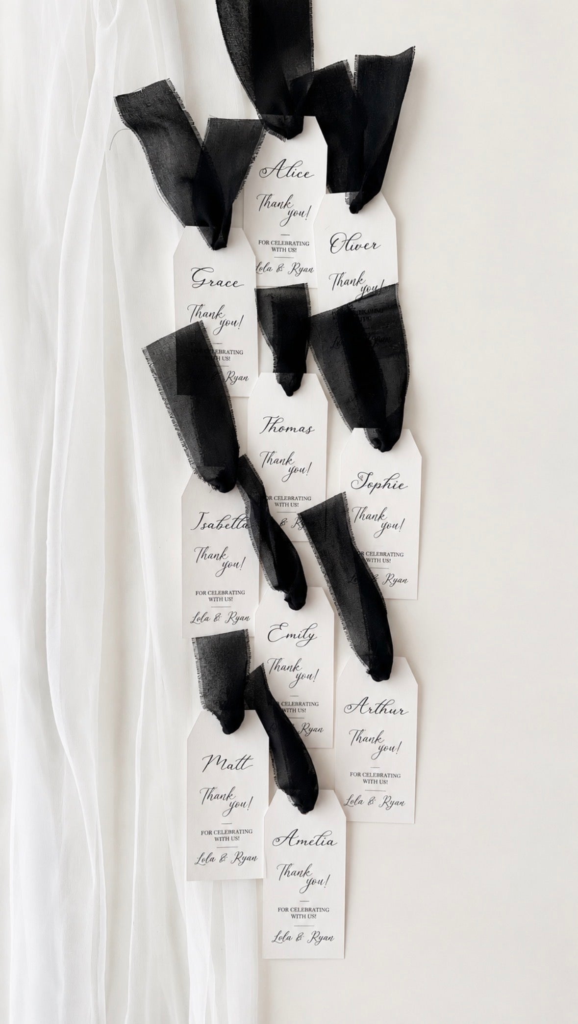 A set of personalised black chiffon ribbon and paper tag favor wraps tied to black ribbon, displayed on a white fabric backdrop. Each name is hand written in black in. 