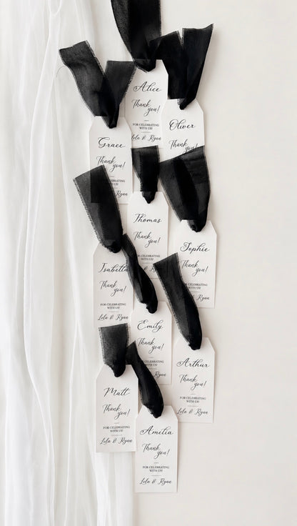 A set of personalised black chiffon ribbon and paper tag favor wraps tied to black ribbon, displayed on a white fabric backdrop. Each name is hand written in black in. 