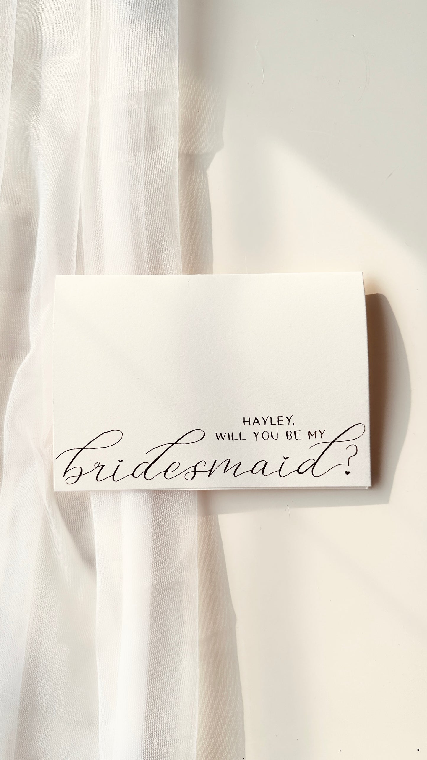 handwritten on cream card  asking ‘Hayley will you be my bridesmaid’ in a mixture of script and standard lettering in black ink