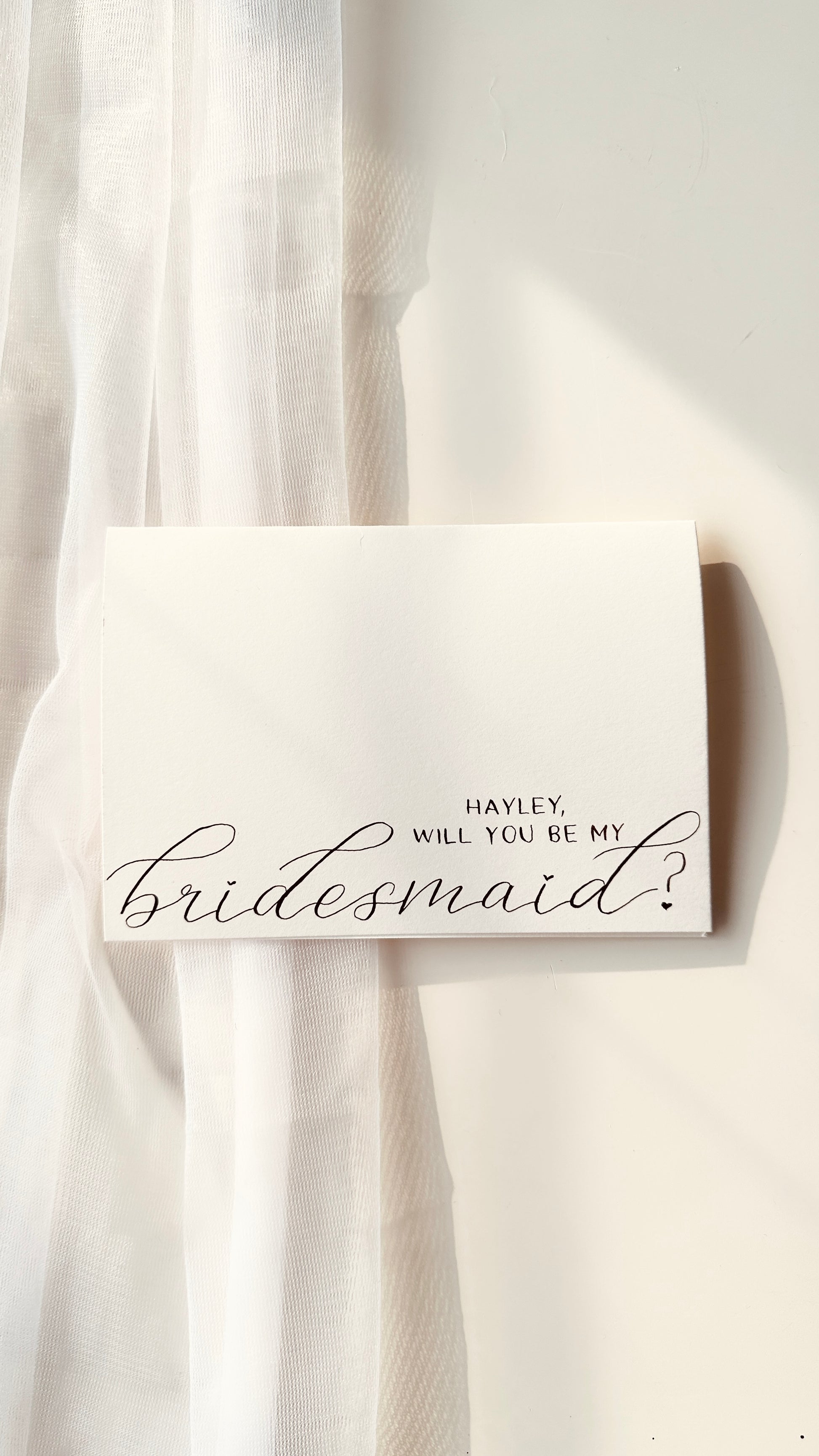handwritten on cream card  asking ‘Hayley will you be my bridesmaid’ in a mixture of script and standard lettering in black ink