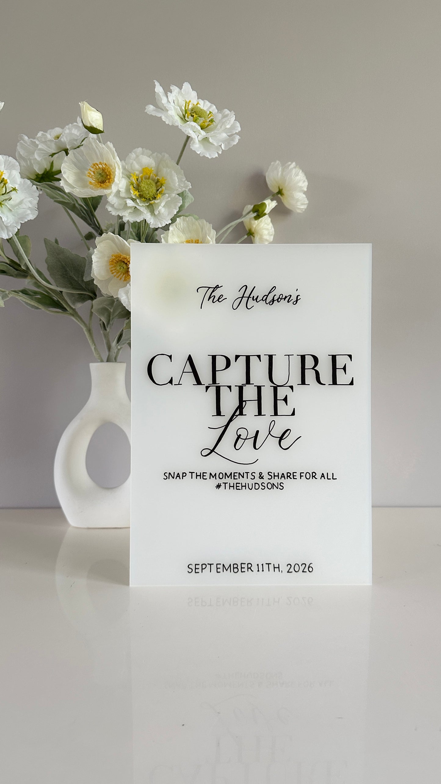 White frosted acrylic with 'Capture the Love' text and flowers in a vase on a light gray background. Sign reads “The Hudson’s, capture the love, snap the moments and share for all #thehudsons, September 11th 2026”