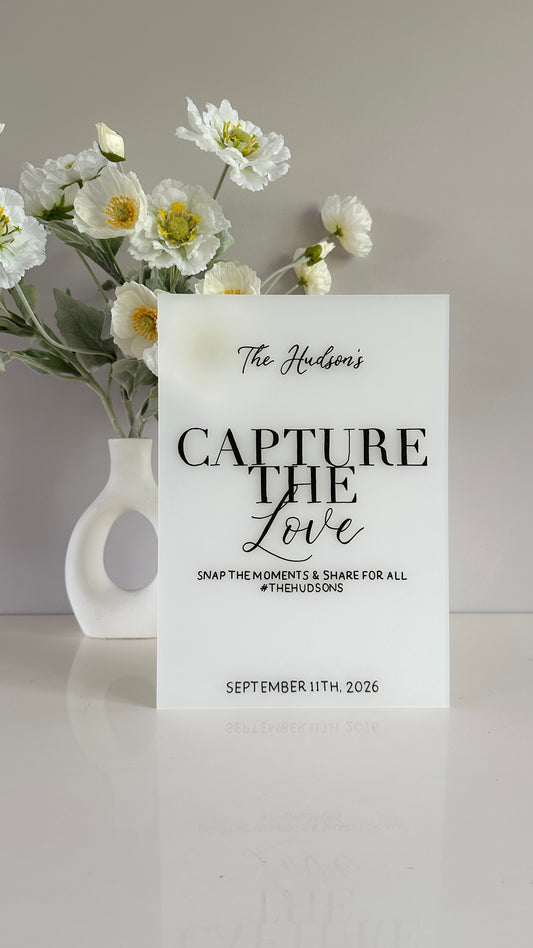 White frosted acrylic with 'Capture the Love' text and flowers in a vase on a light gray background. Sign reads “The Hudson’s, capture the love, snap the moments and share for all #thehudsons, September 11th 2026”