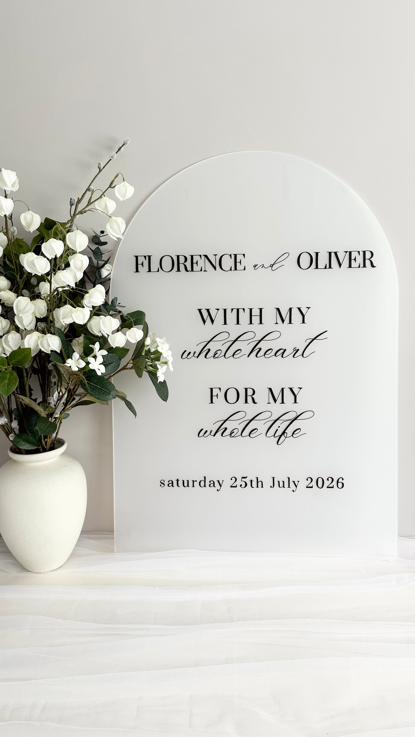 A1 frosted white acrylic with blank ink reading ‘Florence & Oliver, with my whole heart for my whole life, Saturday 25th July 2026’ next to a white and green floral arrangement