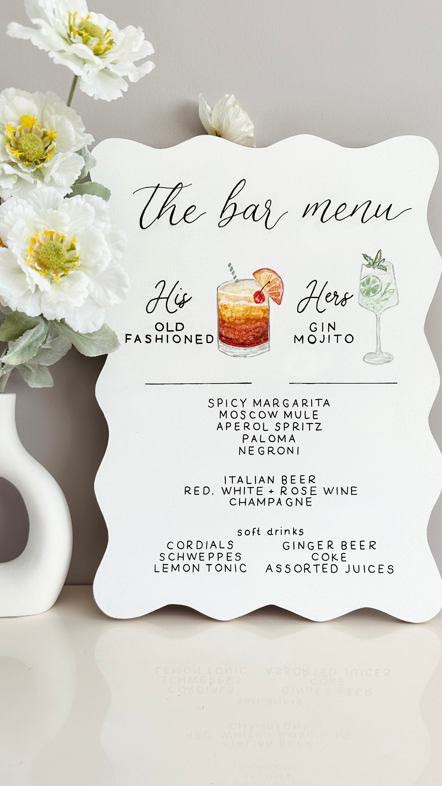 Want A4 Bar menu with hand illustrated cocktail options for 'His' and 'Her' displayed on a white surface with flowers. Writing is black showing cocktail names and bar menu for other drinks available. Cocktails displayed are an old fashioned and gin mojito
