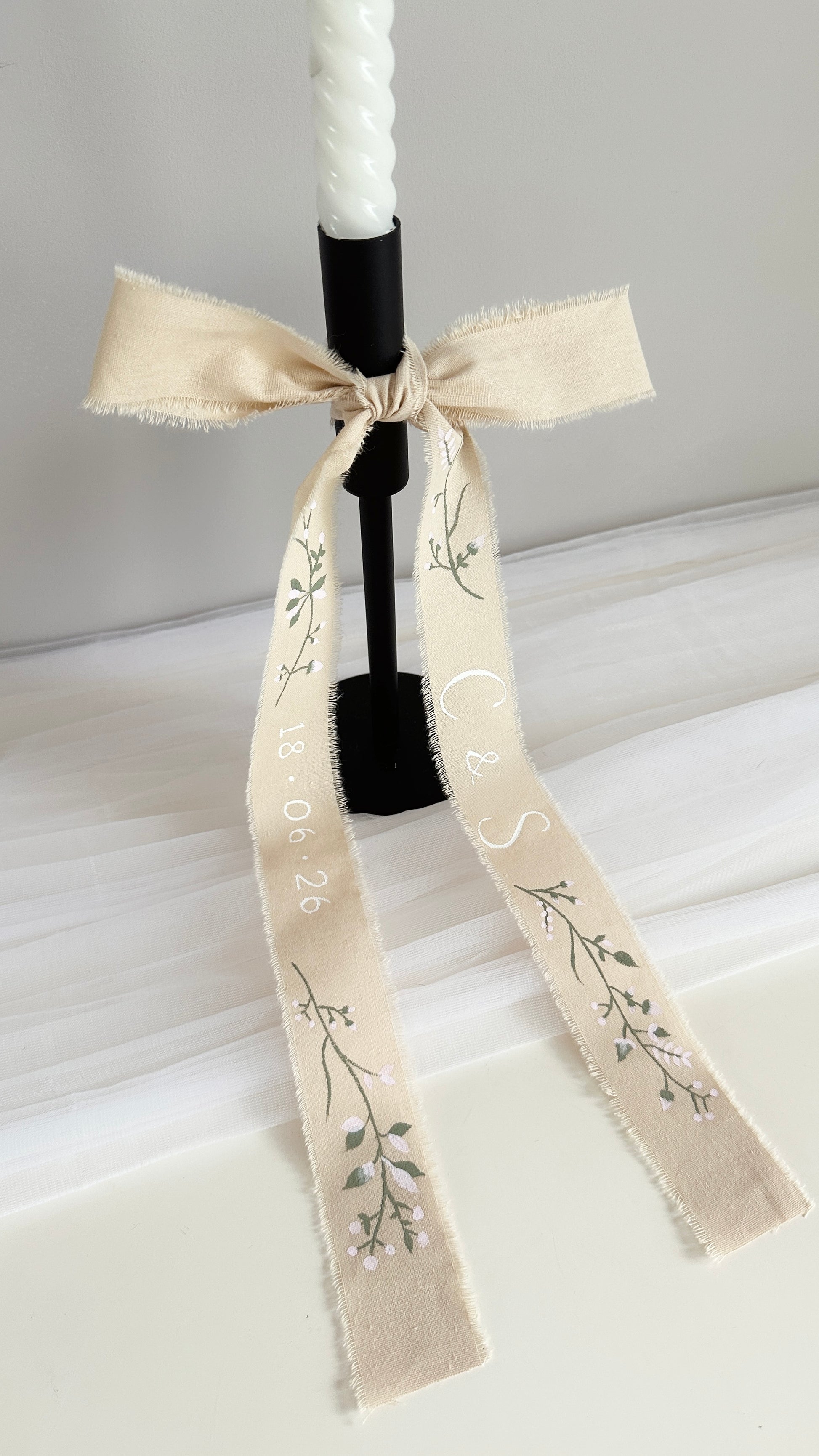 A white taper candle in a black holder, decorated with a cream-colored fabric ribbon tied in a bow. The ribbon features delicate florals hand-painted in sage green and blush pink with the couples initials and dates hand-painted on each side