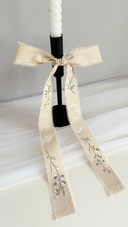 A white taper candle in a black holder, decorated with a cream-colored fabric ribbon tied in a bow. The ribbon features delicate florals hand-painted in sage green and blush pink with the couples initials and dates hand-painted on each side