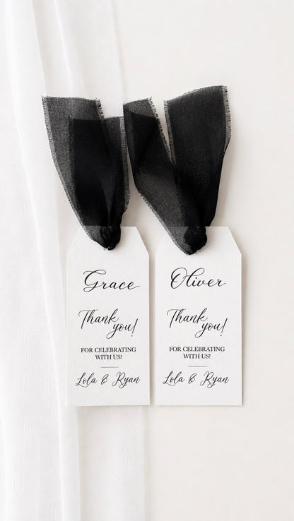 Two white personalised party favor tags with black ribbon bows, handwritten with names and thank you messages, hanging against a white curtain background.