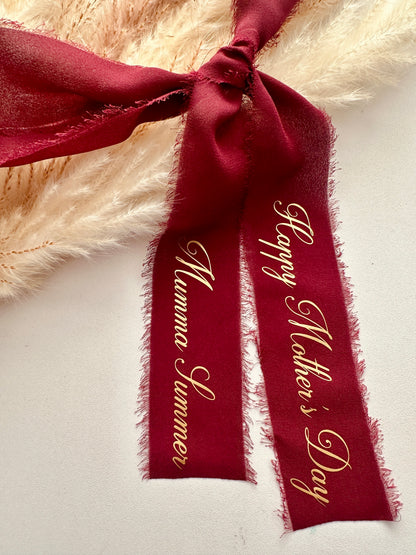 A luxurious deep burgundy chiffon ribbon with delicate frayed edges is elegantly tied into a bow, featuring gold script that reads "Happy Mother's Day" and "Mumma Summer." The ribbon rests on a soft, neutral surface with beige pampas grass in the background, creating a warm and sophisticated presentation.