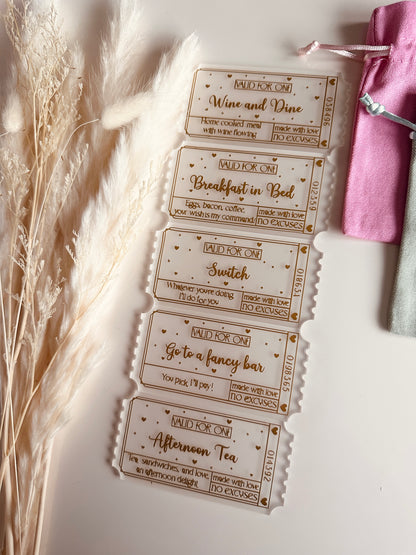 A set of elegant frosted acrylic love coupons with gold engraved text, featuring romantic date ideas like "Wine and Dine" and "Go to a Fancy Bar," displayed next to dried pampas grass and soft pastel pouches.