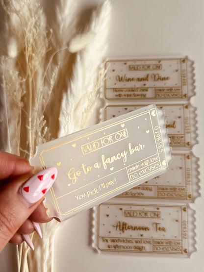 A close-up of a luxurious acrylic coupon reading "Go to a Fancy Bar", held by a hand with heart-themed nail art, against a cozy aesthetic background.