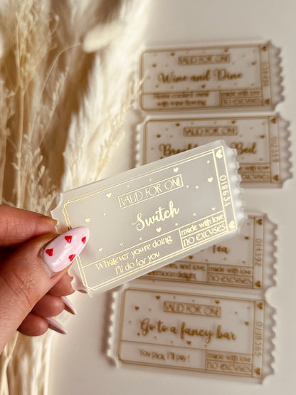 A close-up of a luxurious acrylic coupon reading "Switch", held by a hand with heart-themed nail art, against a cozy aesthetic background.