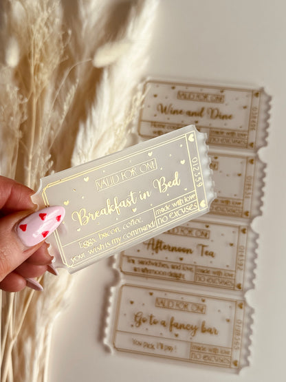 A close-up of a luxurious acrylic coupon reading "Breakfast in bed", held by a hand with heart-themed nail art, against a cozy aesthetic background.
