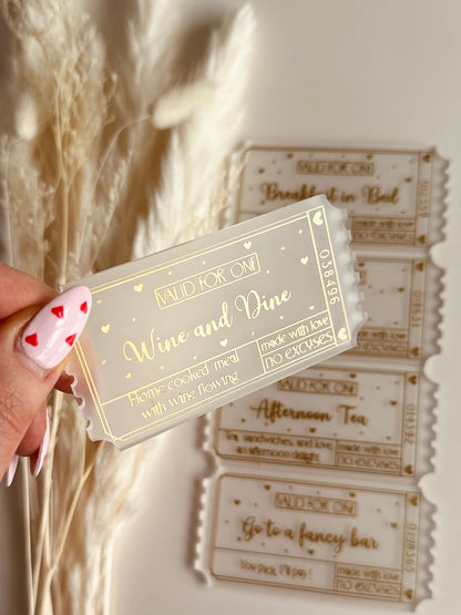A close-up of a luxurious acrylic coupon reading "Wine and dine", held by a hand with heart-themed nail art, against a cozy aesthetic background.
