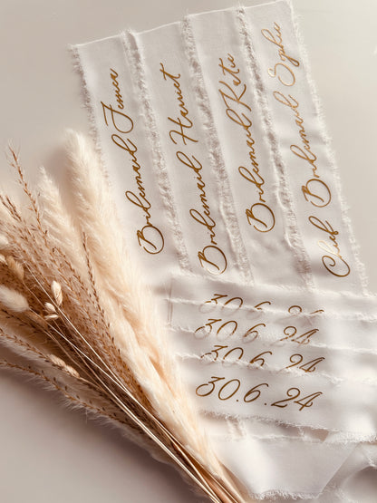 Frayed-edge white chiffon ribbon with calligraphy text, styled with soft neutral pampas grass on a light background, perfect for wedding or event decor. Each bridesmaid names is printed on in gold writing along with the date of the wedding