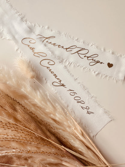 Custom white chiffon wedding ribbons featuring elegant gold calligraphy with couple’s names and wedding date, paired with pampas grass for boho wedding decor.