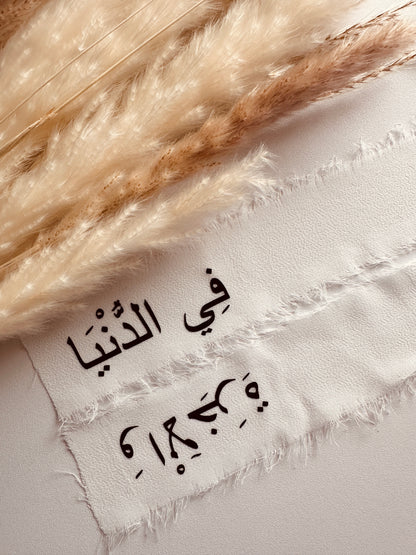 Frayed-edge white chiffon ribbon with Arabic calligraphy text, styled with soft neutral pampas grass on a light background, perfect for wedding or event decor.