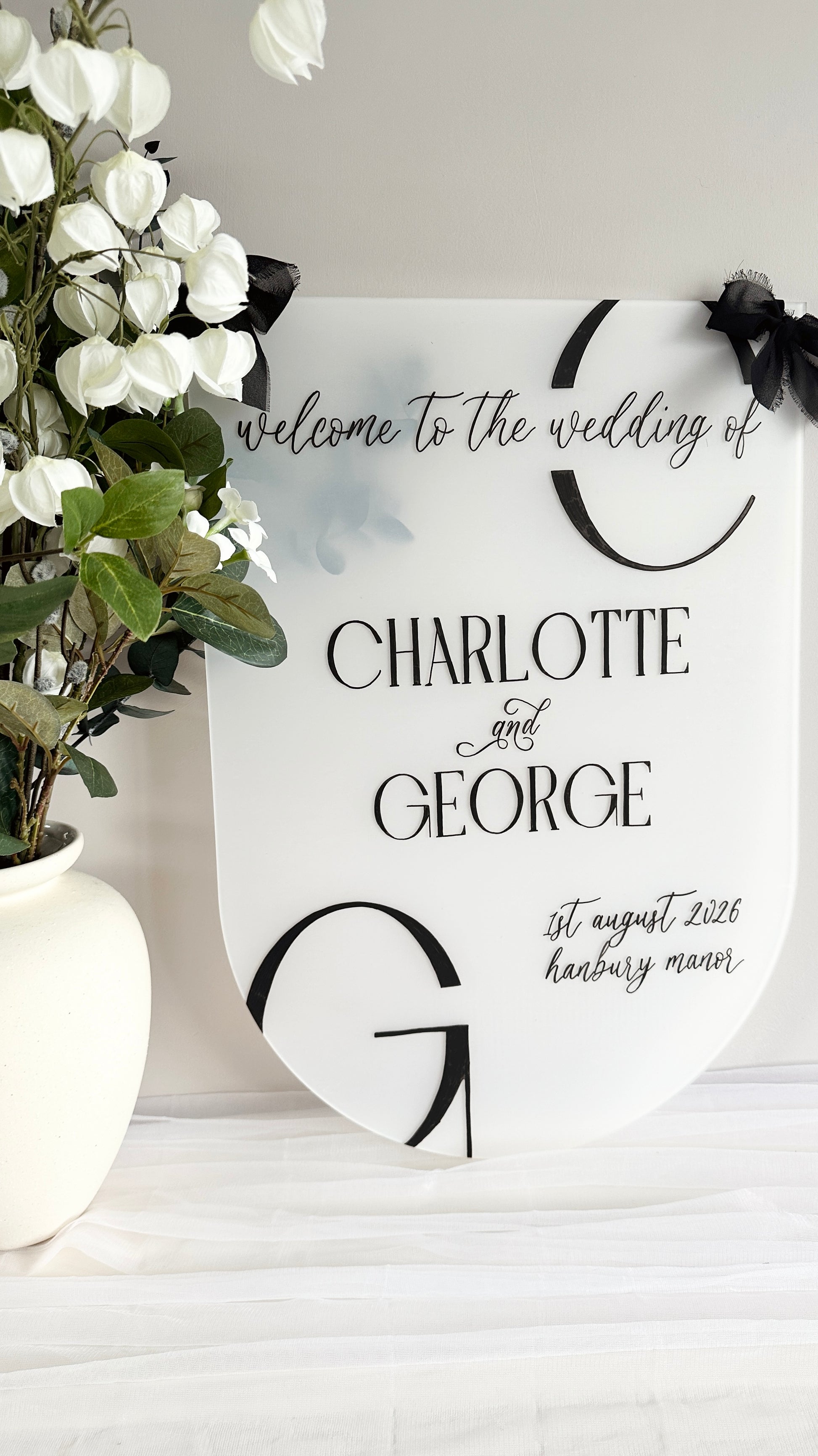 Wedding welcome sign with 'Charlotte and George' on a frosted acrylic sign finished with chiffon black bows