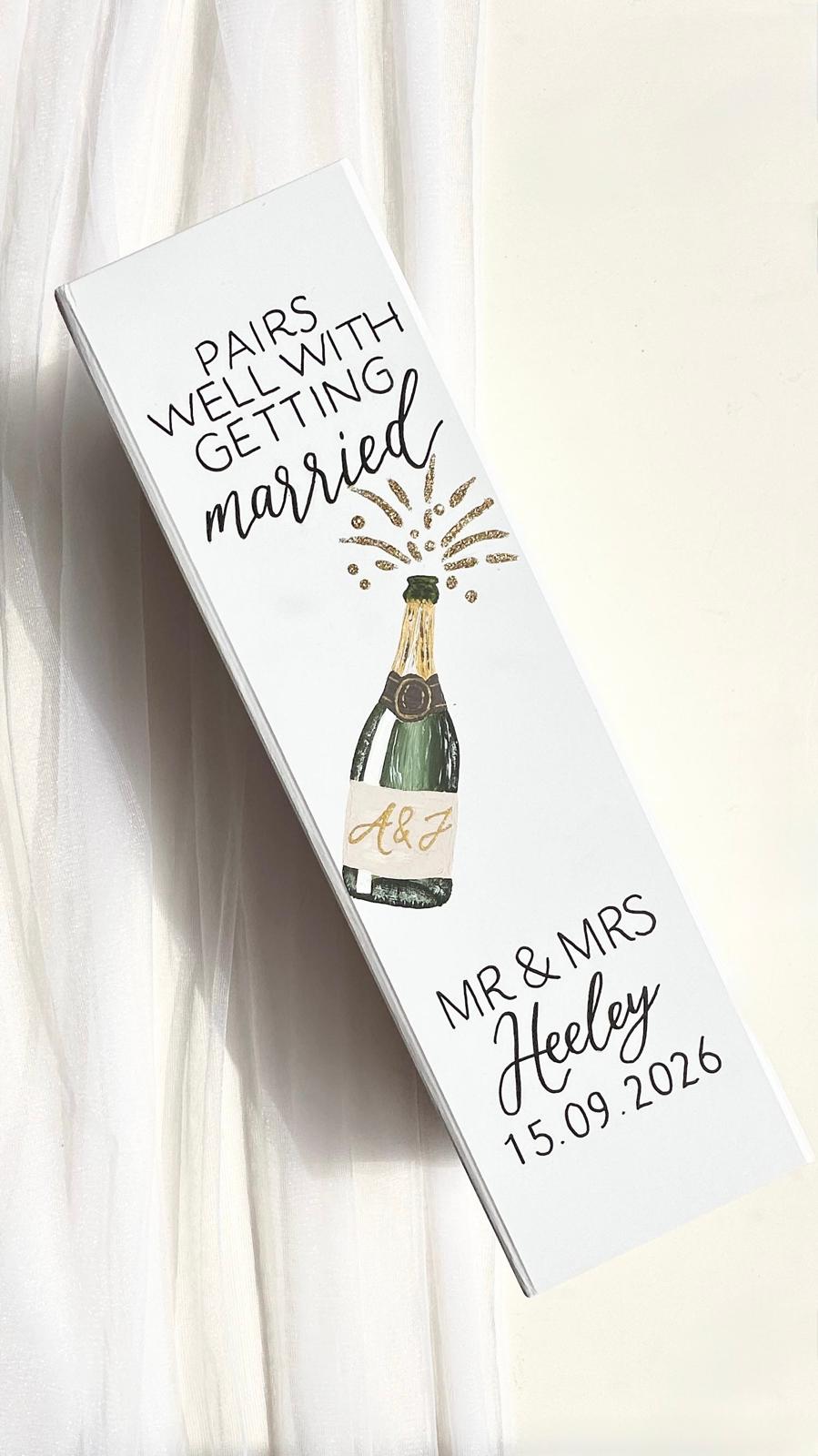 A white rectangular personalised wine gift box with hand painted champagne bottle with couples initial on the label and hand written lettering that reads 'Pairs with getting married' and 'Mr & Mrs Heeley 15.09.
