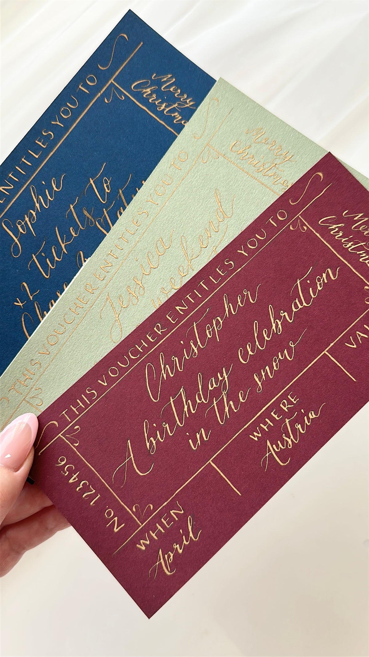 Three Christmas-themed gift voucher cards in navy blue, sage green, and burgundy, each with elegant gold cursive writing, held by a hand against a soft cream fabric background.