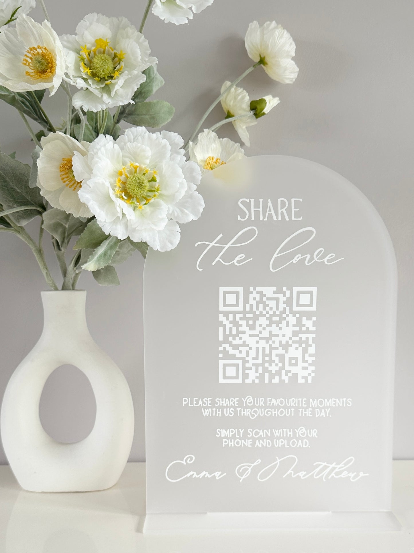 Frosted clear acrylic with white writing and vinyl with QR code that’s scannable via phone for people to share photos of your big day. Writing prompts people to follow the QR code with the couples name 