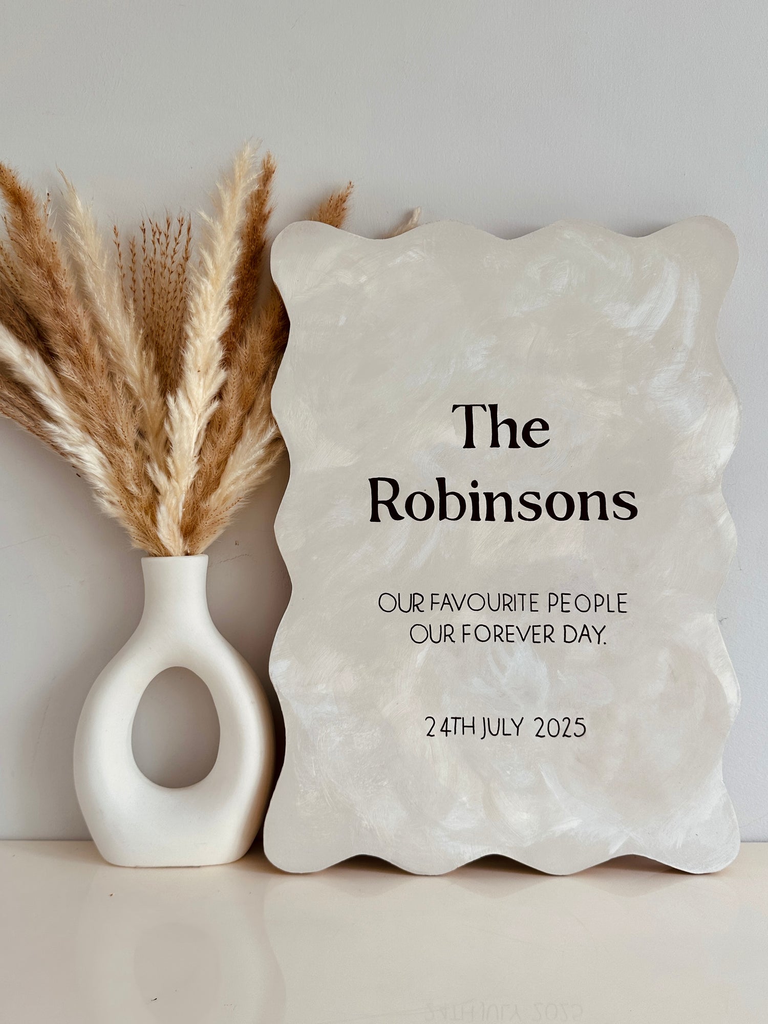 Neutral lime-wash wooden wedding sign with wavy edge and custom text. Decorative stone plaque with personalised text next to a vase with dried plants on a neutral background