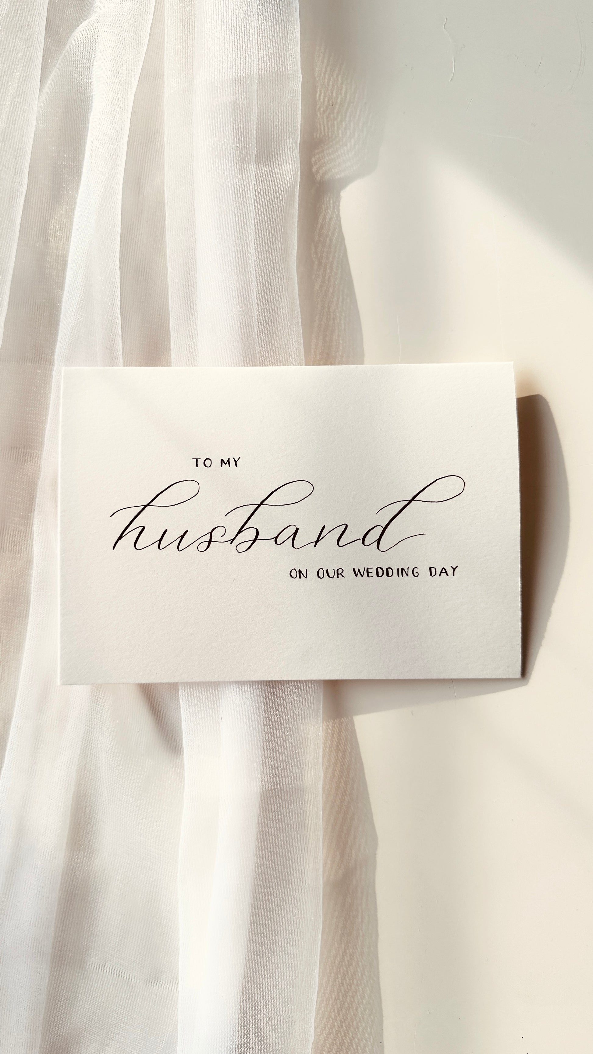 cream premium card with handwritten lettering in black ink reading ‘to my husband on our wedding day’ husband written in script and other words in standing block thin lettering