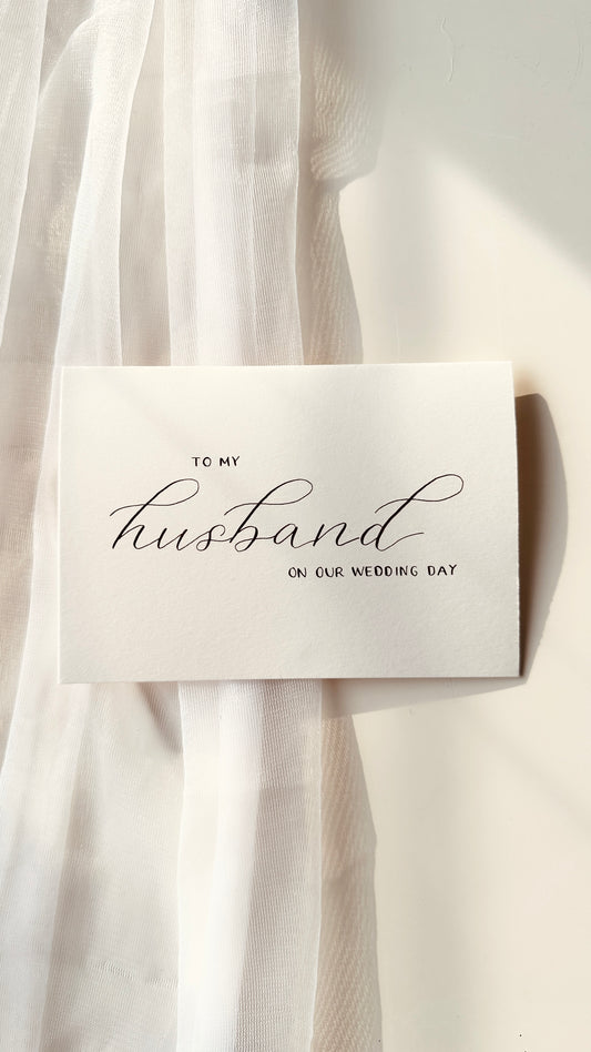 cream premium card with handwritten lettering in black ink reading ‘to my husband on our wedding day’ husband written in script and other words in standing block thin lettering