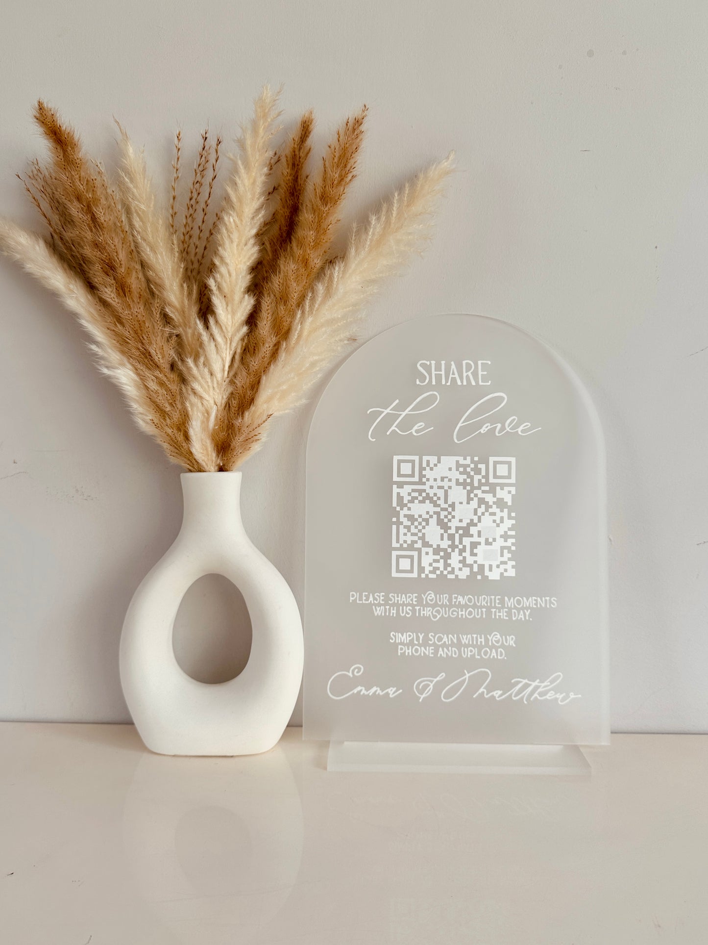 White vase with dried pampas grass and a clear acrylic arch with QR code on with white writing saying 'share the love, please share your favourite moments with us throughout the day. Simply scan with your phone and upload' finished with couples names at the bottom.