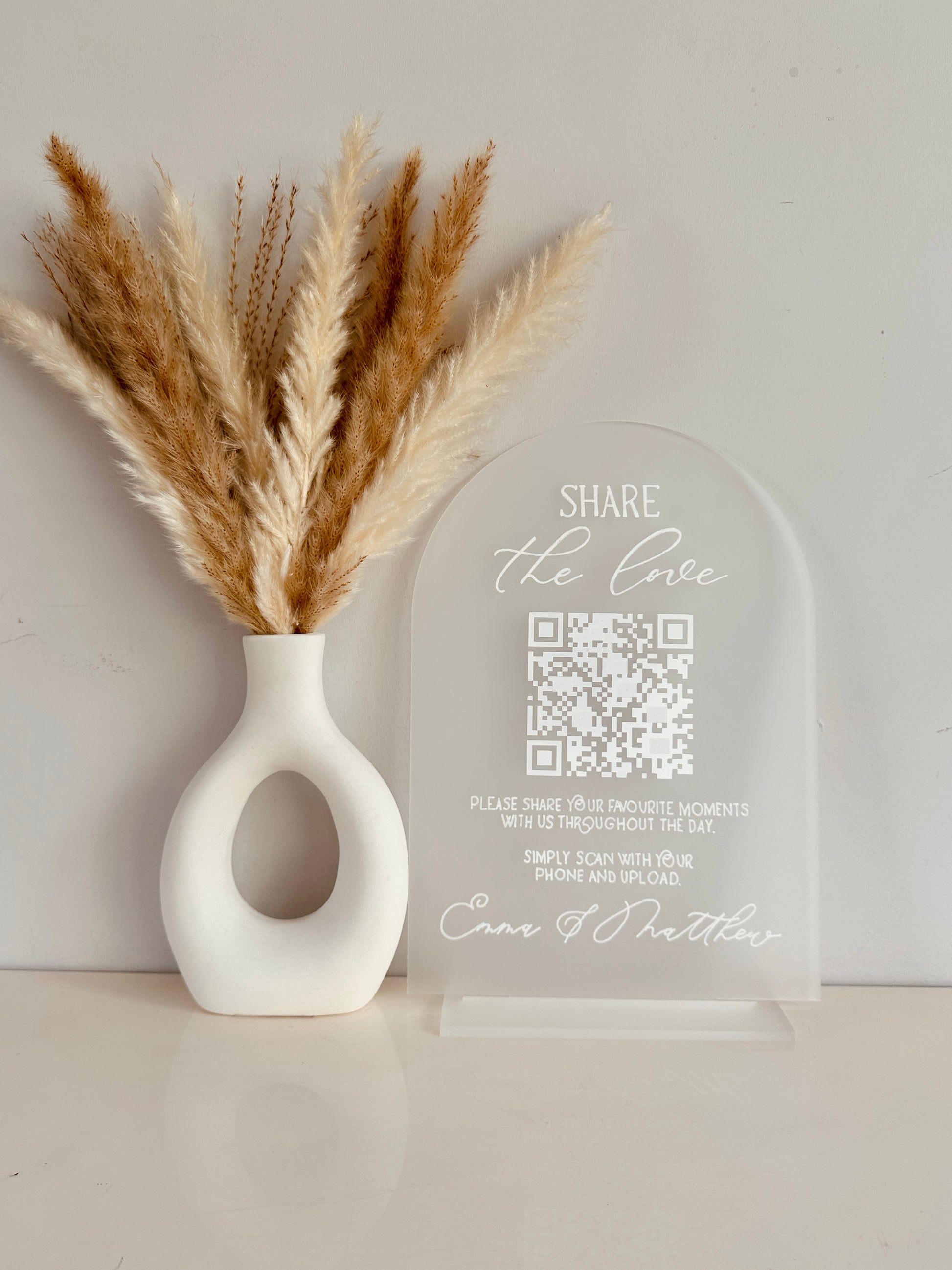 White vase with dried pampas grass and a clear acrylic arch with QR code on with white writing saying 'share the love, please share your favourite moments with us throughout the day. Simply scan with your phone and upload' finished with couples names at the bottom.