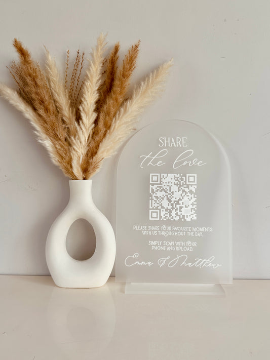 White vase with dried pampas grass and a clear acrylic arch with QR code on with white writing saying 'share the love, please share your favourite moments with us throughout the day. Simply scan with your phone and upload' finished with couples names at the bottom.