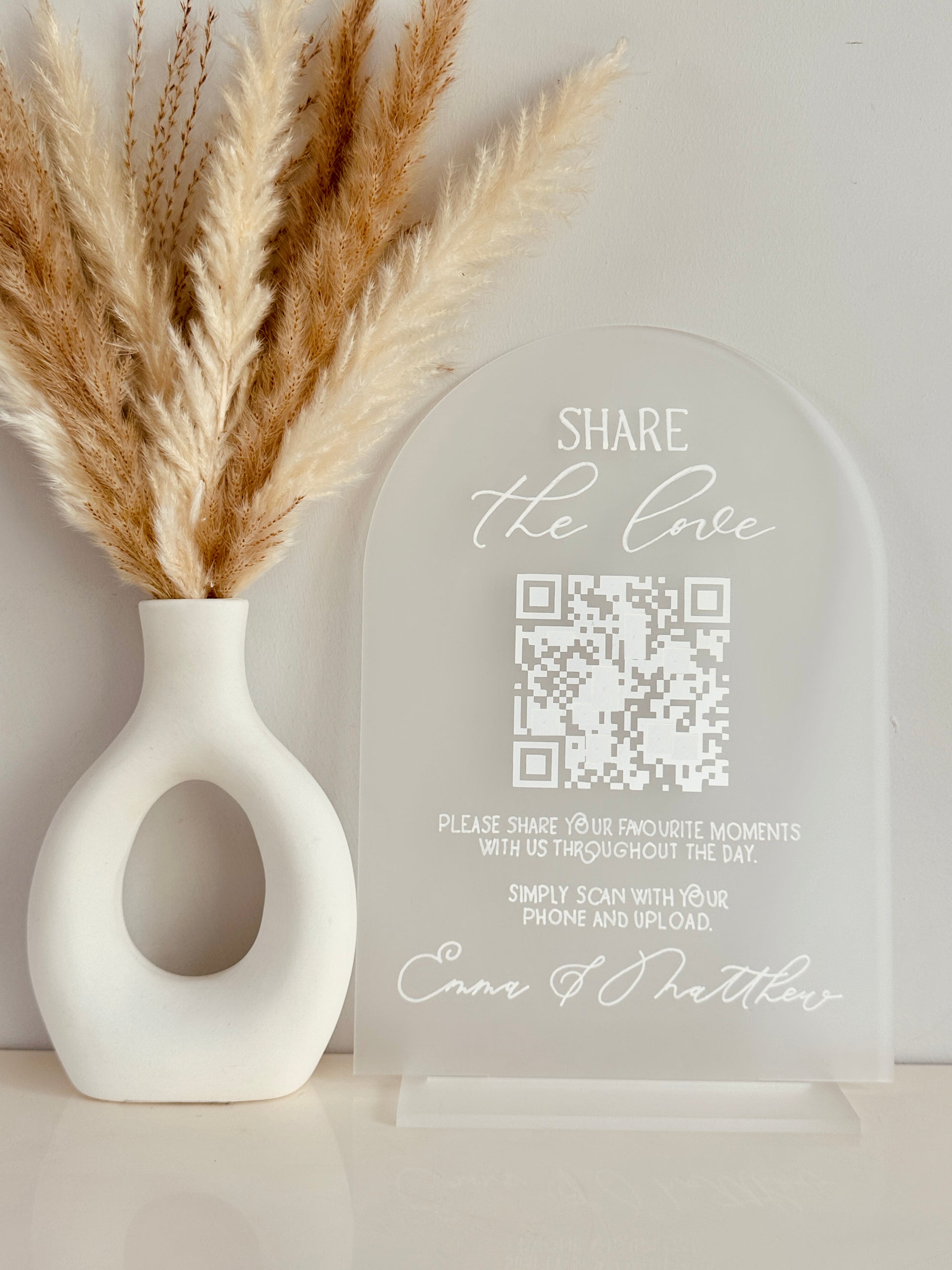 White vase with dried pampas grass and a clear acrylic arch with QR code on with white writing saying 'share the love, please share your favourite moments with us throughout the day. Simply scan with your phone and upload' finished with couples names at the bottom.